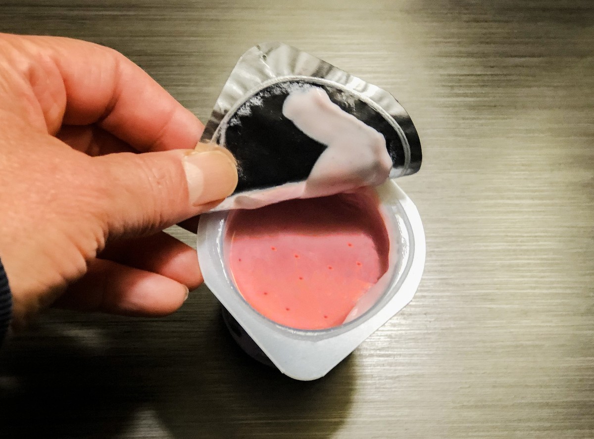 A BPA-free plastic container with strawberry yogurt. A hand is opening the foil wrap lid of the container. There is copy space to the right of the image.