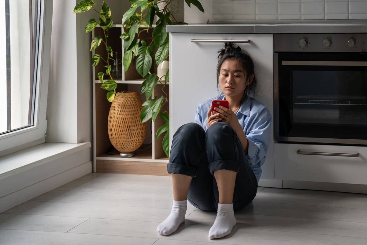 Upset gloomy young Asian woman looking at mobile phone screen feeling sad while sitting on floor Upset gloomy young Asian woman holding cellphone, looking at mobile phone screen feeling sad. Concerned ...