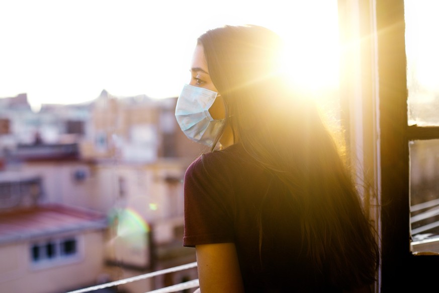 teenage girl wearing protective mask, looking at the city from the window during coronavirus qurantine in Barcelona. Nice sunset with the sun shining in the sky