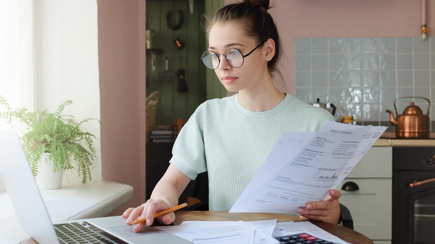 Young woman managing domestic budget, sitting at kitchen table with open laptop, documents and calculator, using touchpad, making notes with pencil