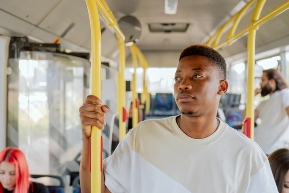 A dark-skinned man stands in the middle of a bus going home, to work, he is tired, he looks out the window, he holds tightly to the railing, a public transport journey with other passengers