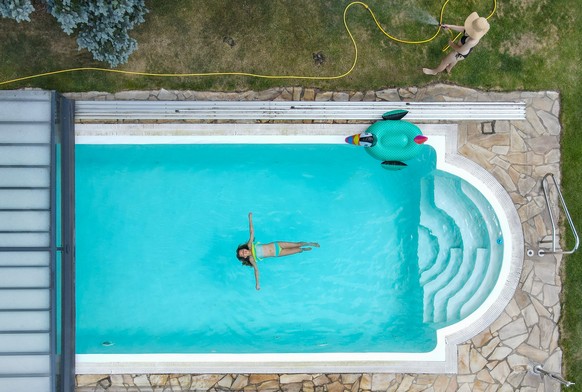 PRODUKTION - 06.07.2022, Brandenburg, Mixdorf: ILLUSTRATION - Ein Kind schwimmt in einem Pool, w�hrend ein anderes M�dchen den Rasen mit einem Wasserschlauch beregnet. (Luftaufnahme mit einer Drohne,  ...