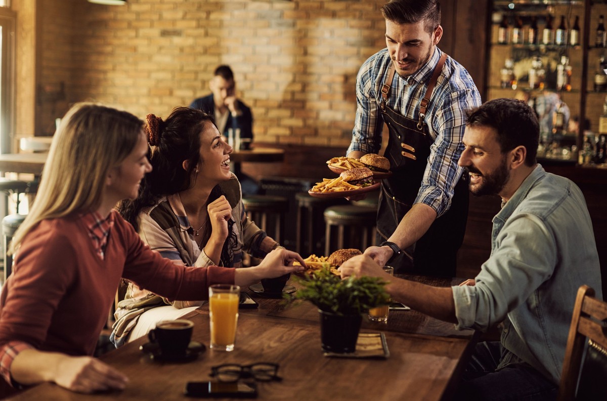 Group of happy friends having fun while waiter is serving them food in a pub.