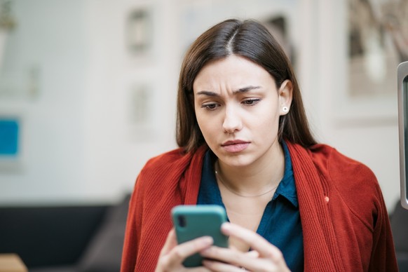 Close up of a depressed Caucasian young woman sitting at home, reading some bad news on her smart phone, feeling sad and worried