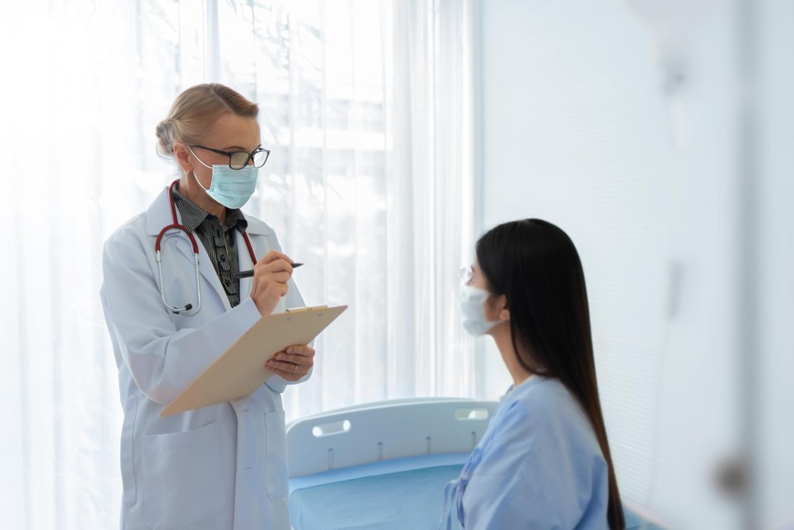 Caucasian Senior female doctor explaining and giving advice to Asian patient in medical room together. Mature physician and Young woman wear protective face masks talking together in hospital.