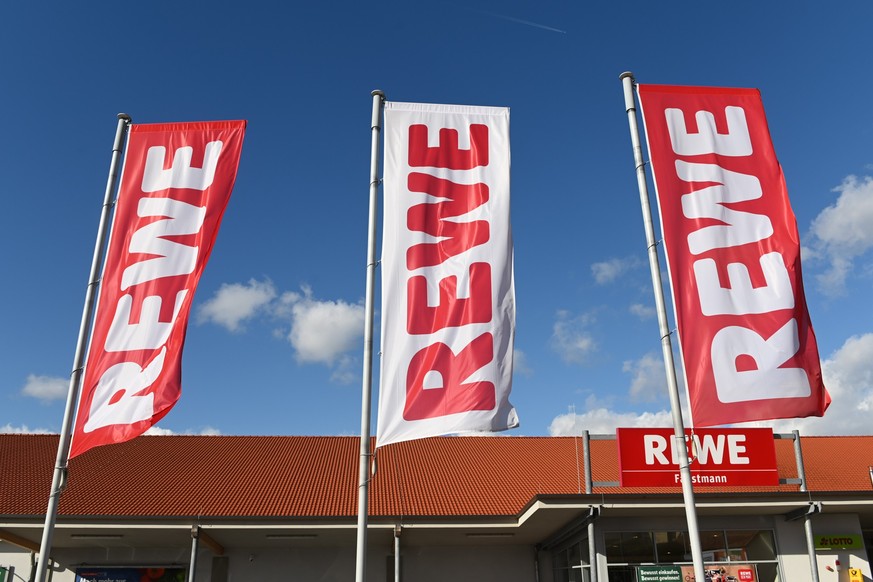 ARCHIV - 25.02.2020, Hessen, Volkmarsen: Fahnen mit dem Logo des Lebensmitteleinzelhändlers Rewe wehen vor einer Filiale. Die Supermarktkette Rewe verzichtet künftig auf ihre Prospekte. (zu dpa «Rewe  ...