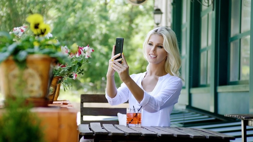 Beautiful young blonde woman in light white shirt relaxing, drinking drinks and taking selfie on smartphone in street summer cafe.