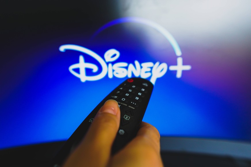 May 10, 2022, Brazil. In this photo illustration a close-up of a hand holding a TV remote control seen in front of the Disney Plus logo. May 10, 2022, Brazil. In this photo illustration a close-up of  ...