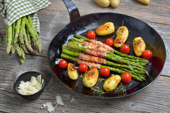 Fried green asparagus wrapped in bacon and served with herb potatoes and tomatoes
with grated parmesan cheese in an old iron frying pan on wooden background