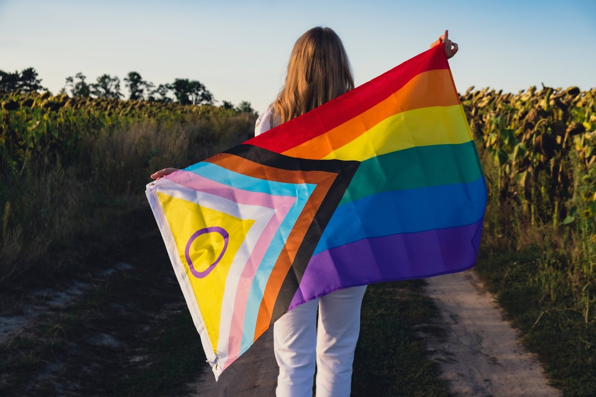 Symbol of LGBTQ pride month. Young woman showing Rainbow LGBTQIA flag waving in wind made from silk material on field background. Equal rights Symbol of LGBTQ pride month. Young woman showing Rainbow  ...