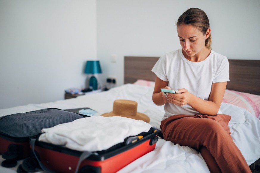 The woman uses a mobile phone after packing her suitcase for travel. She is in her bedroom