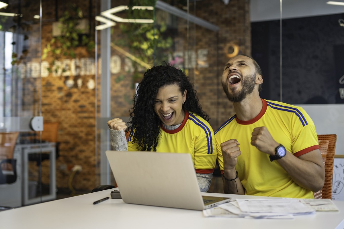 Man and woman excited coworkers watching a colombia national team match on a laptop in the office, while sitting at the boardroom table, businessmen are excited and happy. They are wearing the yellow  ...