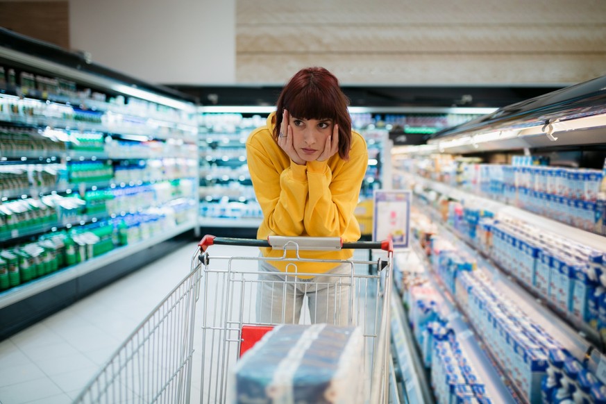 Young woman with red hair buying groceries in a local supermarket, feeling depressed by the high prices in the store and leaning on her shopping cart