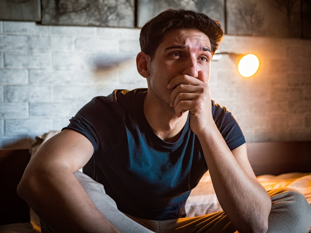 Man covering mouth while sitting on bed crying Young man in domestic clothes sitting on bed against glowing lamp and looking away while crying and covering mouth with hand Copyright: x RECORD DATE NOT ...