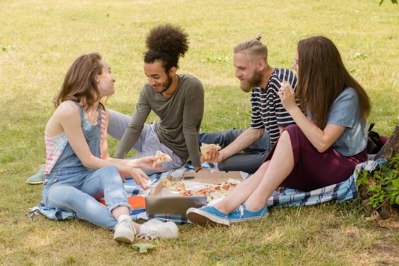 Young multiethnic people sitting on plaid on grass eating. Group cheerful students relaxing having picnic.