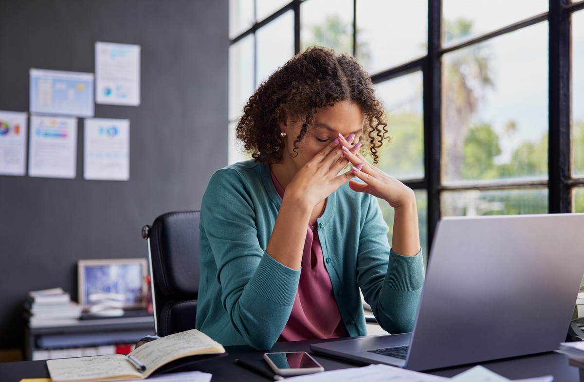 Exhausted businesswoman feeling stressed, holding her head while working on a laptop. Professional african american woman has terrible migraine while managing tasks in office with copy space. Overwhel ...
