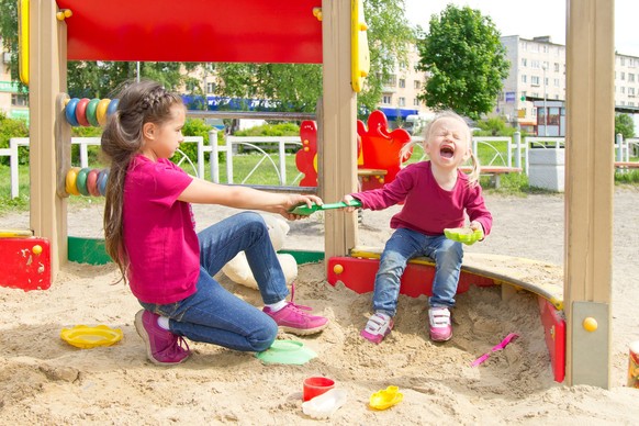 Conflict on the playground. Two sisters fighting over a toy shovel in the sandbox. Kid sister crying all throat