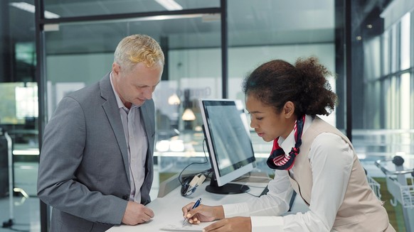 Ein afroamerikanischer Bodenarbeiter einer Fluggesellschaft überreicht einem kaukasischen Geschäftsmannpassagier am Check-in-Schalter des Flughafens eine Bordkarte oder ein Ticket.  Geschäftsreise-Konzept, Modell veröffentlicht, Eigentum veröffentlicht...