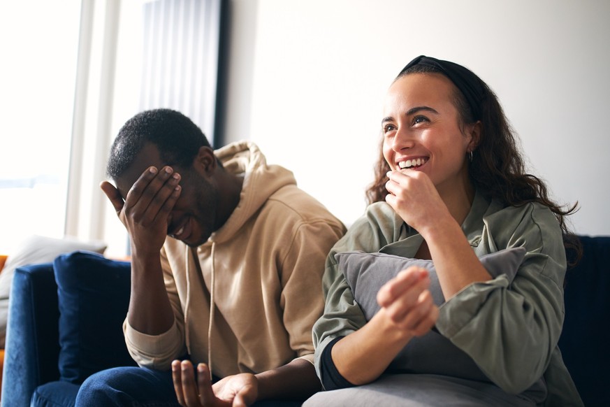 Young mixed ethnicity couple cringing whilst eating popcorn on sofa at home watching or streaming movie on tv together