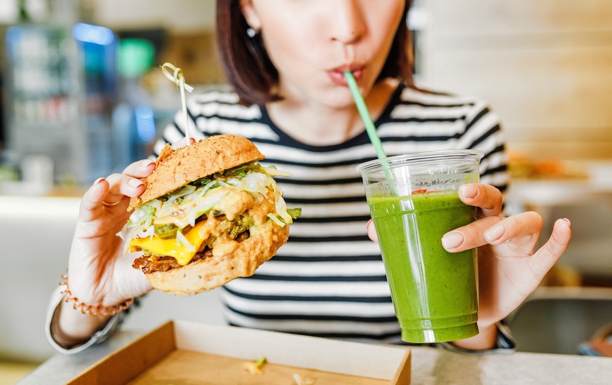 A young woman drinks green smoothies and eats a burger in a vegan fast food restaurant