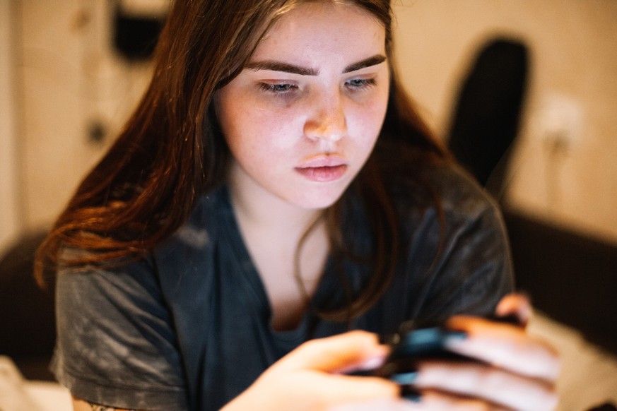 Serious teenage girl using smart phone sitting in bed at home