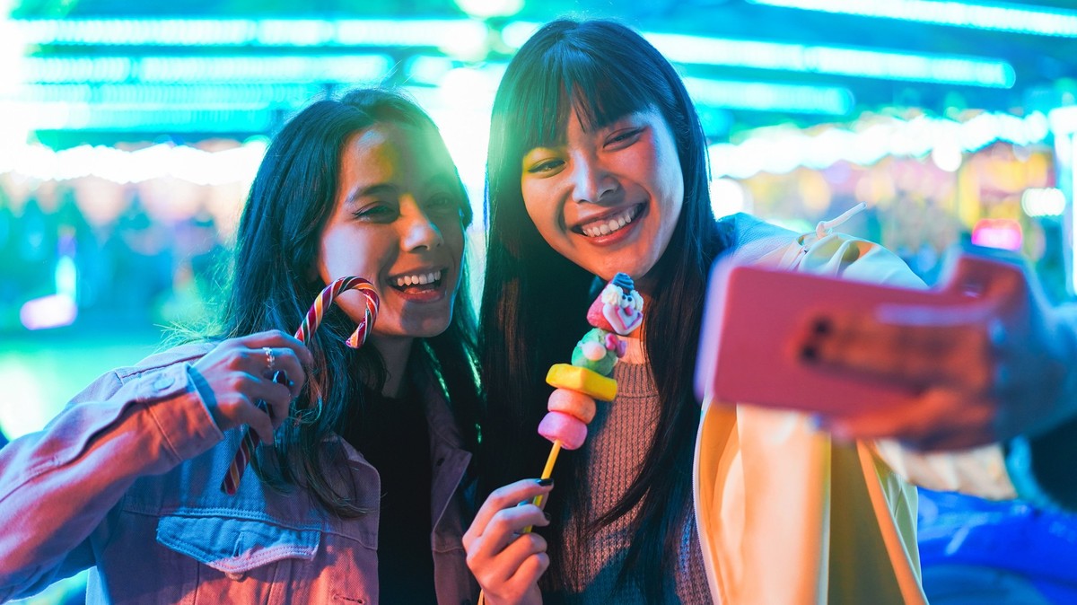 Happy asian girls eating candy sweets and taking selfie at amusement park - Young trendy friends having fun with technology trend - Tech, friendship and influencer concept - Focus on right female face