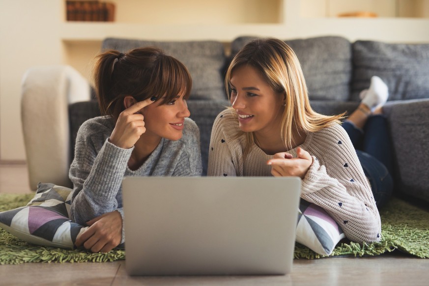 Two young cheerful women are lying in living room carpet and using laptop.