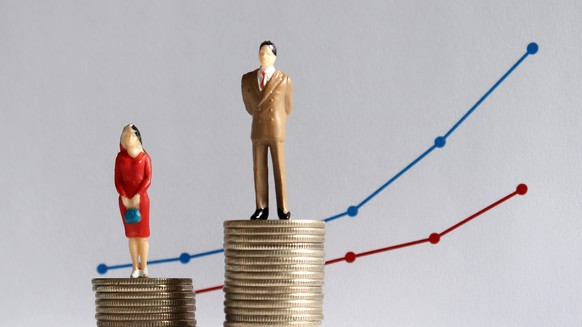 The concept of continuing gender pay gap. A miniature man and a miniature woman standing on top of a pile of coins at different heights in front of a bar graph.