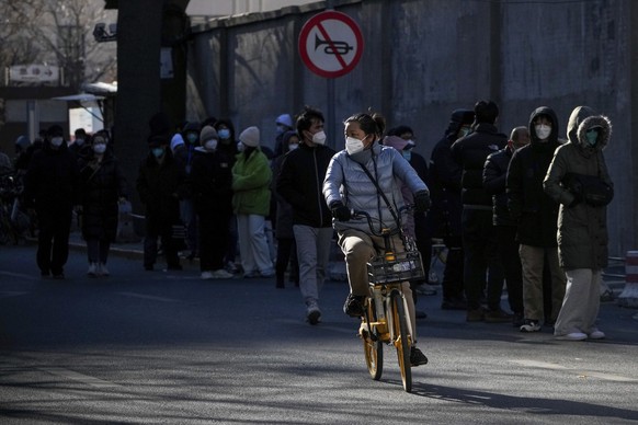 A masked woman bicycles past residents lining up to enter the fever clinic of a hospital in Beijing, Sunday, Dec. 11, 2022. Facing a surge in COVID-19 cases, China is setting up more intensive care fa ...