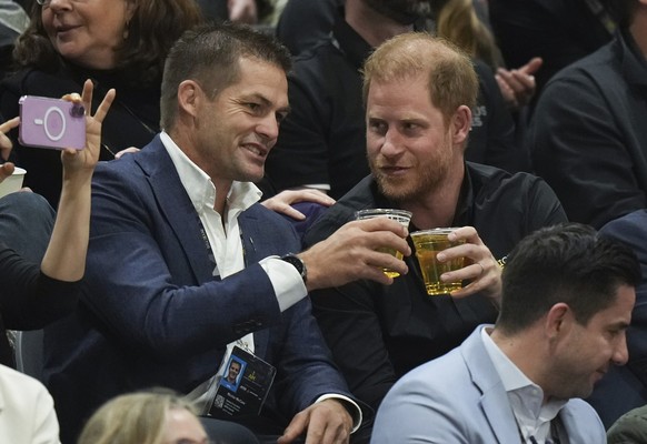 13.02.2025, Kanada, VANCOUVER: Der britische Prinz Harry (r), Herzog von Sussex, trinkt ein Bier mit dem pensionierten neuseeländischen Rugbyspieler Richie McCaw, während er das Spiel zwischen den USA ...