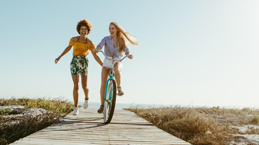 Excited woman riding bike down the boardwalk with her friends running along. Two female friends having a great time on their vacation. Panoramic shot with lots of copy space on background.