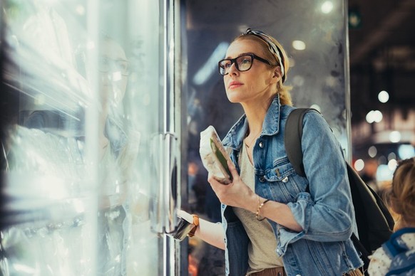 Close up shot of a beautiful blonde woman with blonde hair tied in a bun, wearing black glasses. She opened a fridge and is looking inside choosing products to buy.
