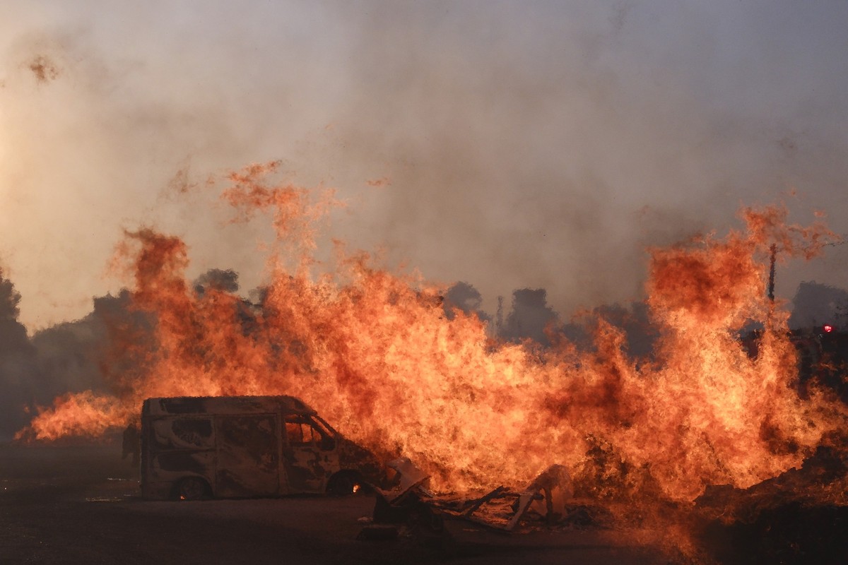 12.08.2024, Griechenland, Athen: Ein großes Feuer verbrennt ein Fahrzeug im Norden von Athen, während Hunderte von Feuerwehrleuten gegen einen großen Waldbrand vorgehen, der am Rande der griechischen  ...