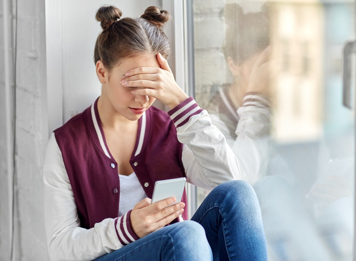 technology, cyberbullying and people concept - sad teenage girl with smartphone sitting on window sill. teenage girl with smartphone sitting on windowsill ,model released, Symbolfoto