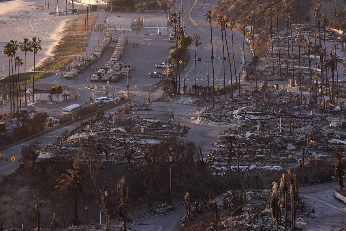 12.01.2025, USA, Los Angeles: Eine ausgebrannte Nachbarschaft ist entlang des Pacific Coach Highway nach dem Palisades-Feuer zu sehen, während sich mehrere Fahrzeuge der Nationalgarde am Strand aufste ...
