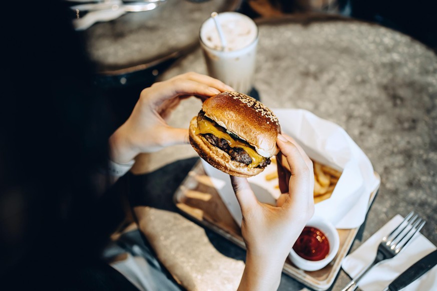 Over the shoulder view of young woman enjoying her meal, eating a freshly made beef and cheese burger with fries and having a glass of iced milkshake in a restaurant