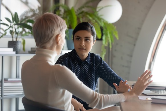 Indian and caucasian businesswomen negotiating sit at desk in office. Lawyer consulting client during formal meeting. Job interview and hiring process. Diverse colleagues discussing project concept