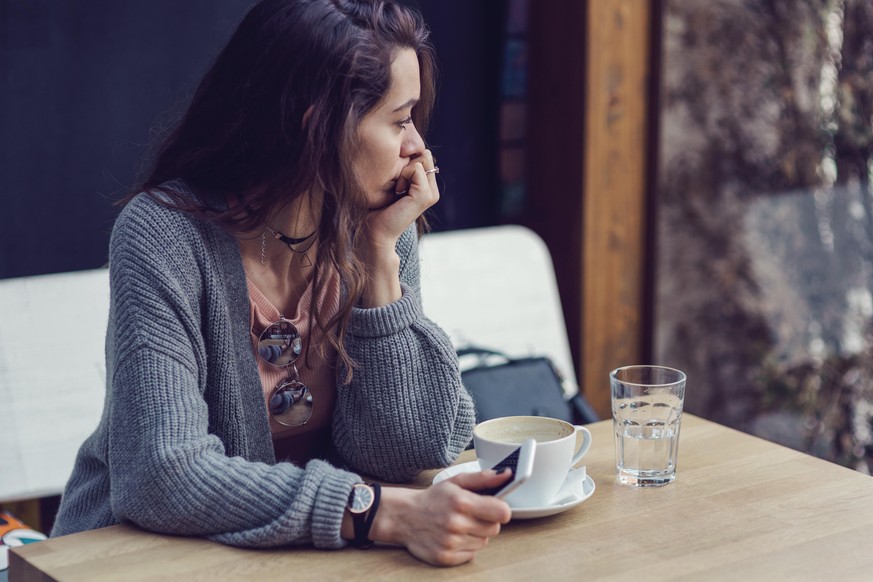 Brunette female holding her mobile phone, looking through the window and drinking coffee