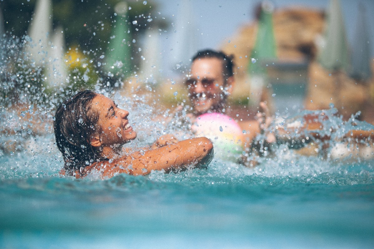 Two people are standing at the pool and have fun together. Girl is in focus and she is trying to catch a colorful ball which guy trow to her. She have big toothy smile and a lot of drops around face.  ...