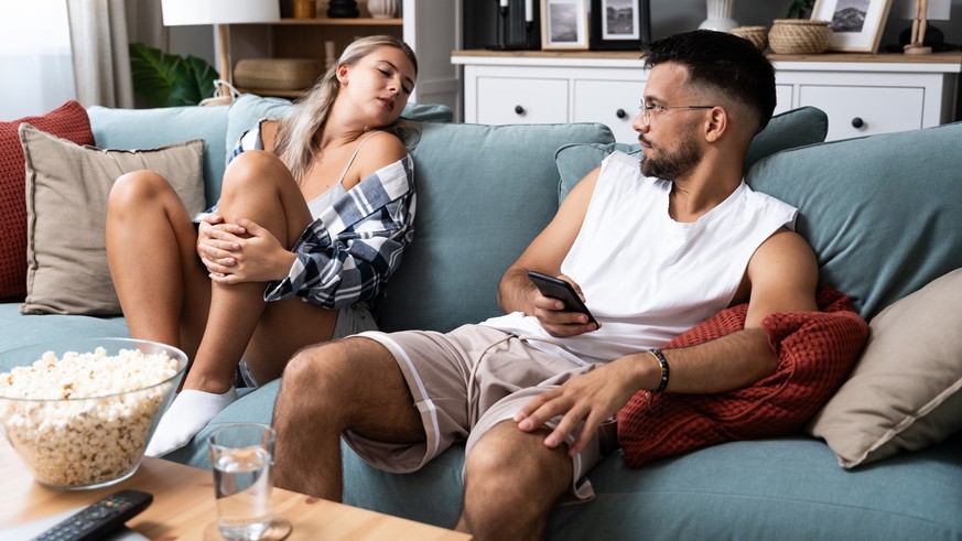 Young boyfriend making fun and jealous his girlfriend pretending that he is texting with other girl on his cell or mobile phone while they sitting on the sofa in their apartment Model Released Propert ...
