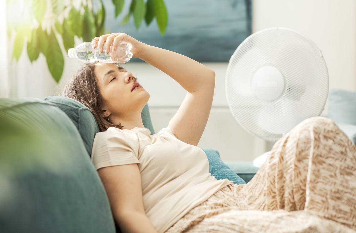 Young woman sitting on the couch at home and keeping a fresh water bottle on her forehead, she is suffering from the heat