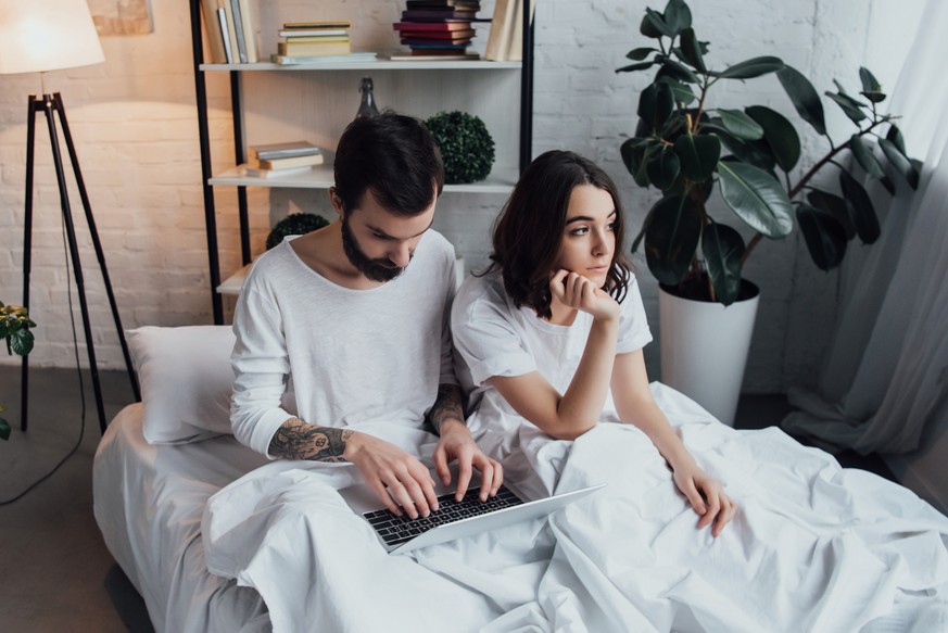 man lying in bed and using laptop while bored woman propping chin and looking away in bedroom