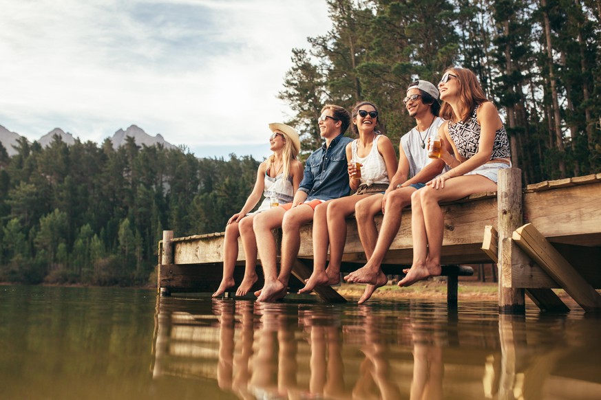 Group of happy young friends having fun and drinking beer while sitting on the jetty at the lake. Young men and woman enjoying a day on Lake.