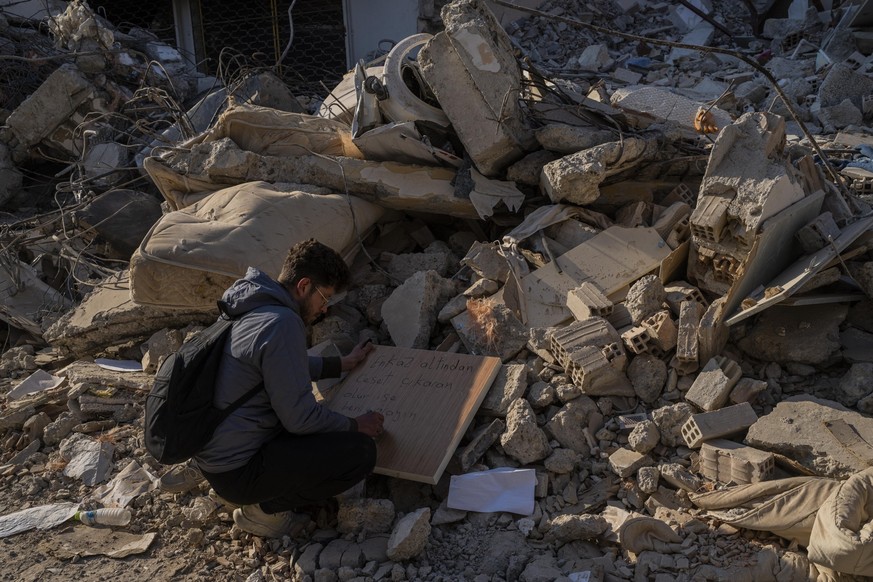 13.02.2023, Türkei, Antakya: Ein Mann schreibt Kontaktinformationen auf ein Brett, für den Fall, dass eine Leiche unter den Trümmern eines zerstörten Gebäudes in Antakya im Südosten der Türkei gefunde ...
