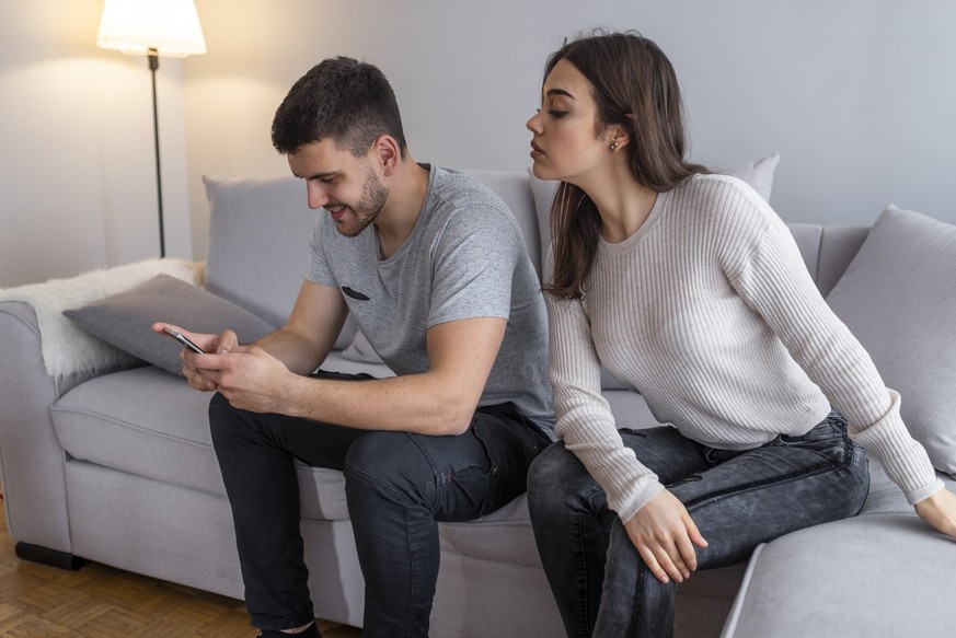 Emotional upset couple sitting on couch and quarreling about smartphone, mistrust concept. Jealous suspicious mad wife arguing with obsessed husband holding phone texting cheating on cellphone
