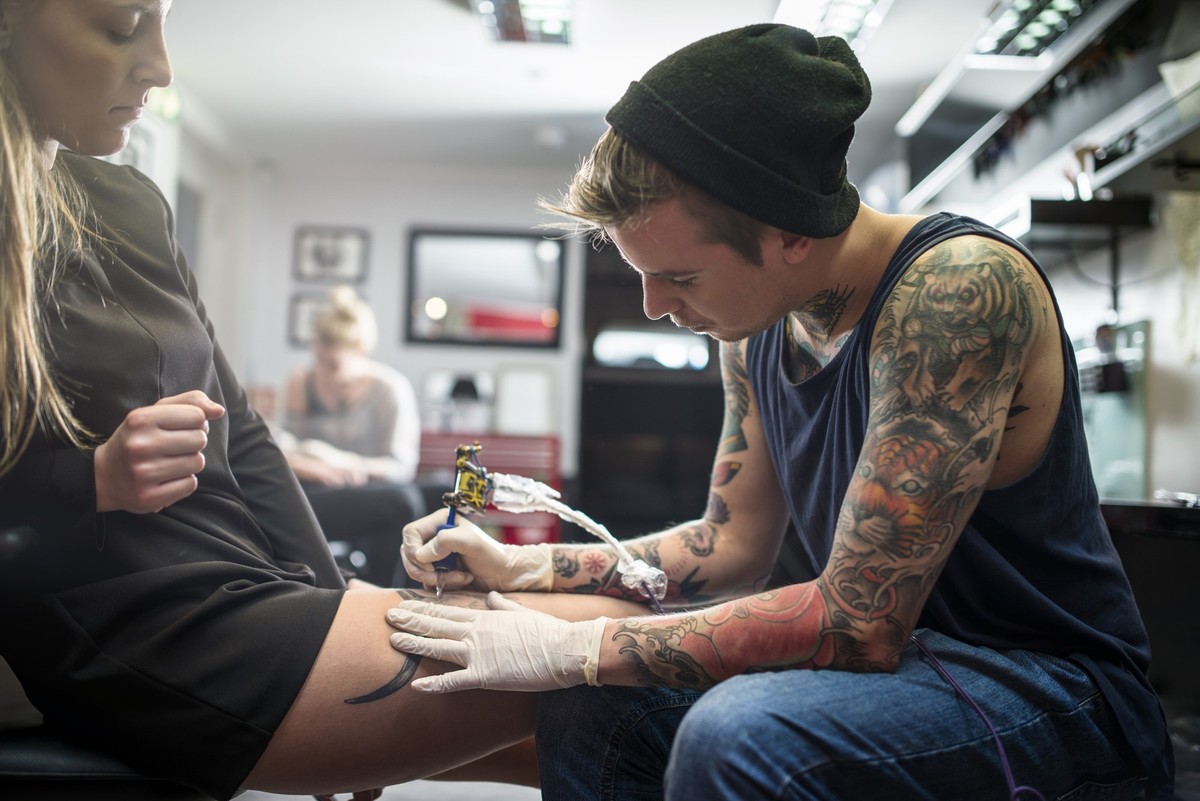 A photo of expert tattooing female customer&#039;s lap. Tattdio. Woman is looking at man making tattoo on her leg.