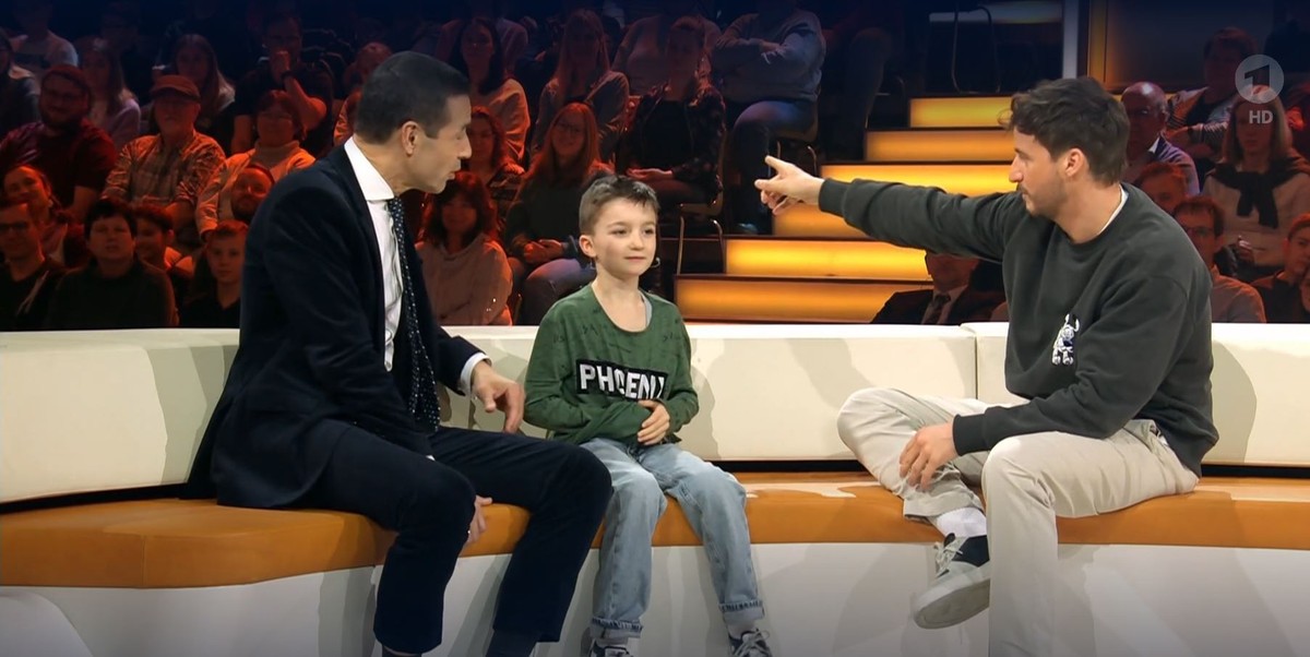 Tommi Schmitt zeigt ins Publikum und suggeriert, dass Jamal Musiala dort sitzt.
