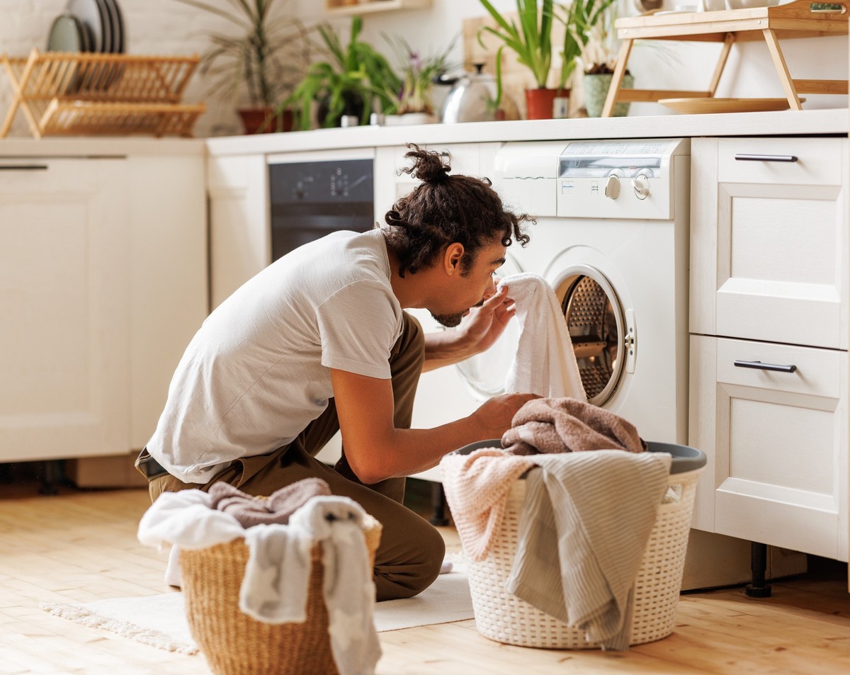 Young ethnic black man householder with curly hair sitting on floor near washing machine while doing laundry in kitchen at home
