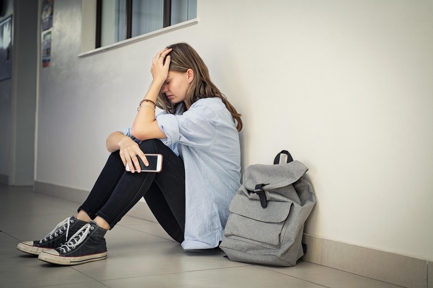 Upset and depressed girl holding smartphone sitting on college campus floor holding head. University sad student suffering from depression sitting on floor at high school. Lonely bullied teen in diffi ...