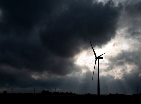 23.07.2023, Schleswig-Holstein, Klanxbüll: Ein Windrad zeichnet sich auf einem Feld vor dunklen Wolken und der Sonne ab. Foto: Daniel Bockwoldt/dpa +++ dpa-Bildfunk +++